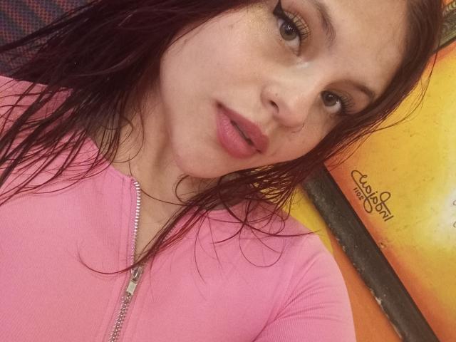 SophieHill with wet brown hair, pink zip-up shirt, and pink lipstick, showing a cute face with slightly parted lips. She has a small nose and long eyelashes.