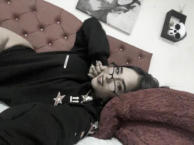 CristalSweety, lying on a bed, wearing glasses, black hoodie with star and lettering, hand near face, smiling, dark hair, pink tufted headboard, red pillow, grayscale wall art.
