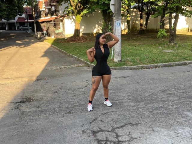 CristalSweety avec une grande poitrine, un débardeur noir ajusté, un short moulant, des baskets blanches, un tatouage sur la cuisse droite, debout sur un trottoir fissuré, une main sur la tête, une journée ensoleillée, des arbres en arrière-plan..