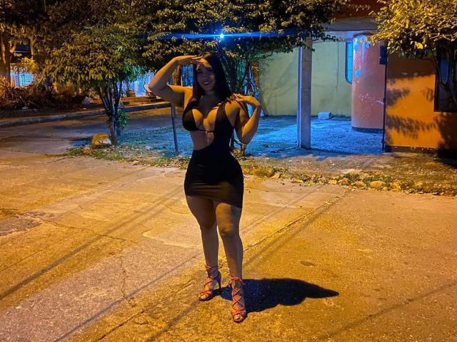 CristalSweety, a curvy woman with big tits and a small ass, wears a black, cut-out dress and red strappy heels, standing on a dimly lit, gravel-covered street at night.