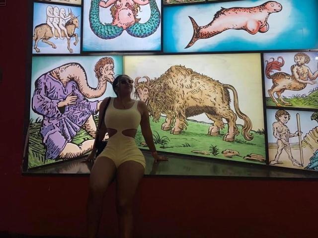 CristalSweety with big ass, small tits, in a white cutout dress, sitting against a wall of colorful, weird animal paintings. She has dark skin, a curvy figure.