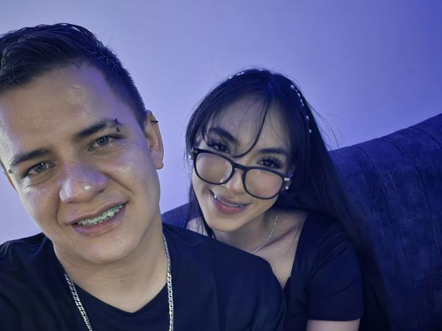 AriandDante with glasses and black hair leaning on a couch, smiling, wearing a black shirt. AriandDante with short black hair, metal chain, and tooth grill, smiling.