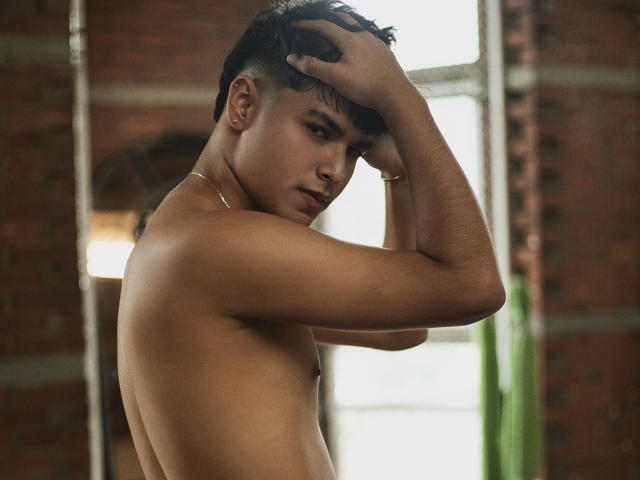 Shirtless MathewAdams with tan skin, dark hair, and a gold necklace, raising his right hand to his head, looking serious. His ass is partially visible, background blurred.