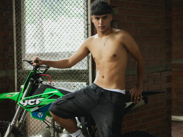 Shirtless MathewAdams with a slim, toned body, black pants, and a flat stomach, leaning on a green Yamaha dirt bike, wearing a black cap, in a brick room.