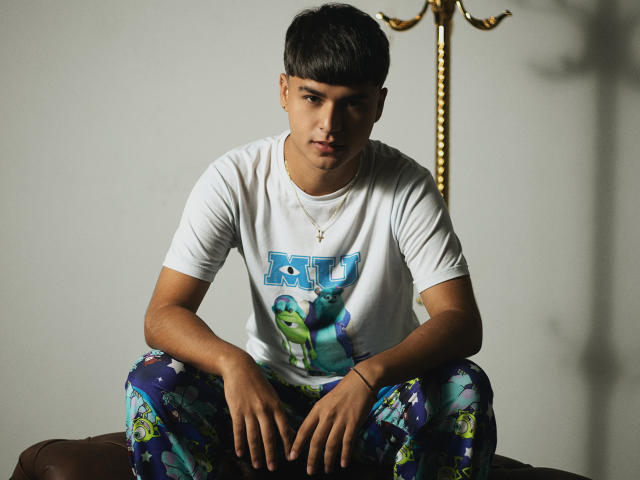 MathewAdams, dark-haired dude, white tee, colorful pants, sitting on brown couch, looking serious, arms resting on knees, gold necklace, shadow on wall, soft lighting, casual style.