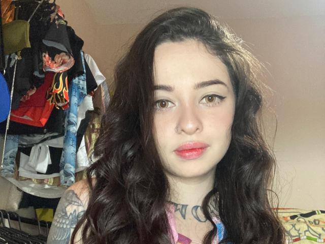 ArtemisSolo has wavy dark brown hair, brown eyes, red lipstick, a nose ring, and a tattoo on her right arm. She's wearing a colorful top, looking at the camera.