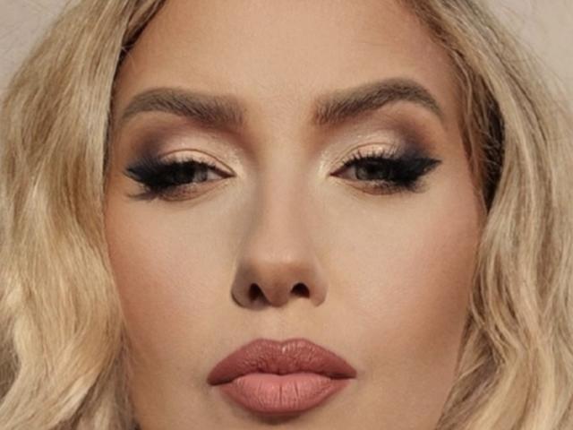 OndinaNoir's face with blonde wavy hair, tan skin, and full makeup including dark eyeliner, mascara, and pinkish-brown lipstick. Her eyes are half-closed and her lips are slightly pursed.