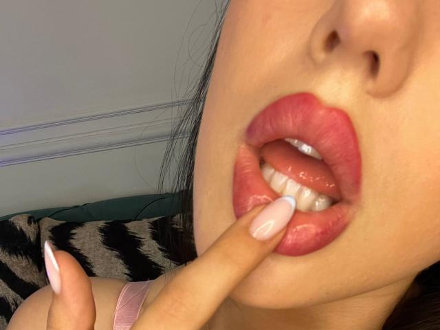 AlenaCapricious's face, slightly open mouth with pink lipstick, teeth visible, tongue partially out, finger with pink nail touching bottom lip, dark hair, fair skin.