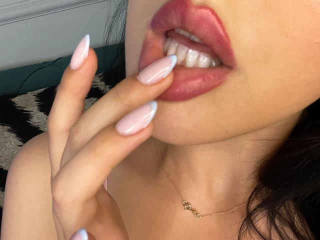 AlenaCapricious's pink glossy lips are slightly parted, showing white teeth, as she touches her upper lip with her long, pink and blue manicured fingers. She wears a gold necklace.