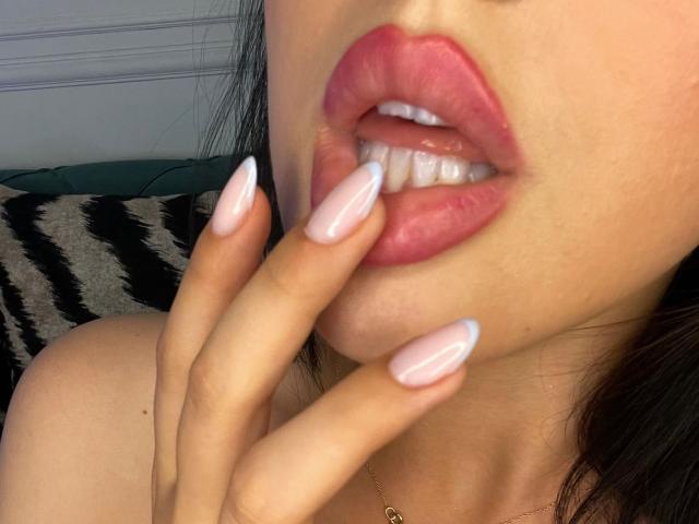 Los labios rosados y brillantes de AlenaCapricious están ligeramente entreabiertos, mostrando los dientes blancos, y sus uñas largas y rosadas la rozan suavemente el labio inferior. Lleva un pequeño collar de oro..