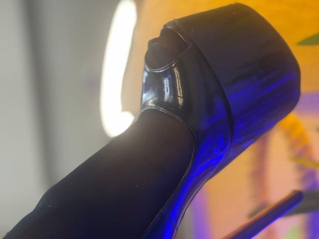 Black high heel shoe with shiny, blue-tinged patent leather, showing the back and heel. The heel is thick and glossy, with a slightly curved, pointed design.