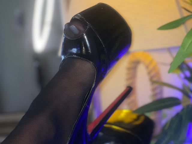 A shiny black high heel with a blue tint, covering AlenaCapricious' foot, featuring a red stiletto heel and sheer black stockings.