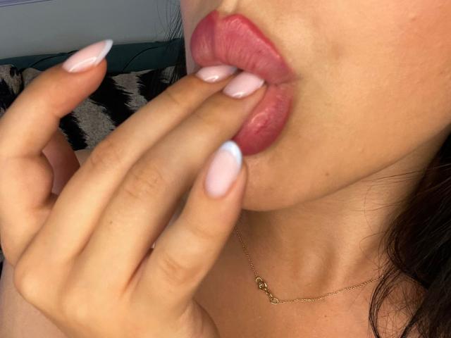 AlenaCapricious with pink glossy lips, fingers with long, pale pink and white nails, slightly parting her lips, touching her lower lip. She's wearing a delicate gold necklace.