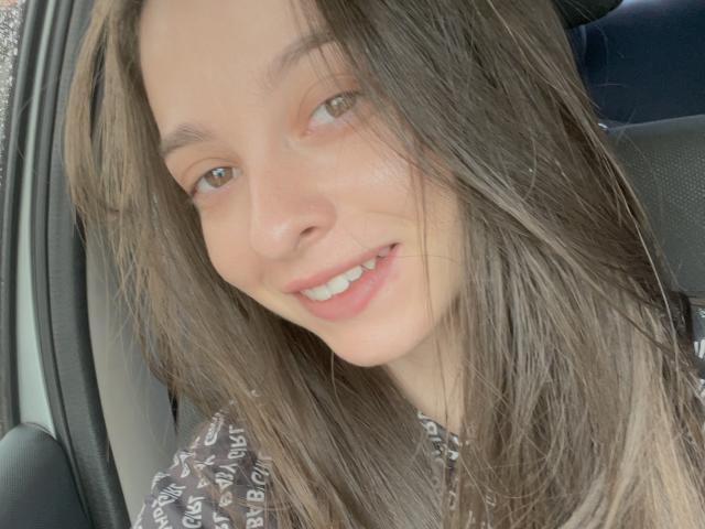 ArtemisSolo's face with long brown hair, green eyes, and a small smile, wearing a brown patterned shirt, sitting in a car. Her tits are not visible.