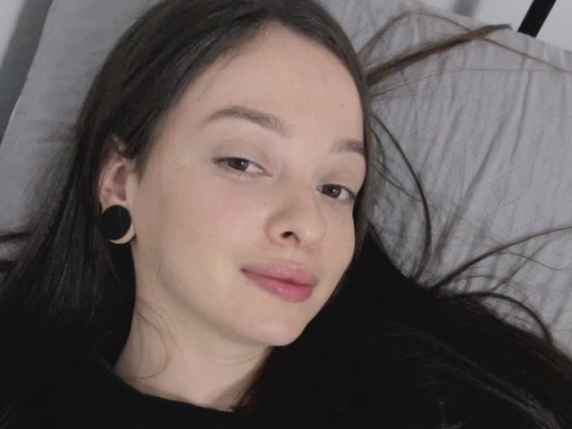 ArtemisSolo has light skin, long brown hair, black ear gauges, and is lying down with a slight smile, showing pink lips. Her brown eyes are half-open.