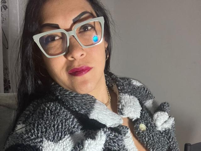 MellissaCortez with black hair, wearing gray and white fuzzy coat, teal glasses with blue dot, and red lipstick, looking slightly to the side. Boobs not visible.