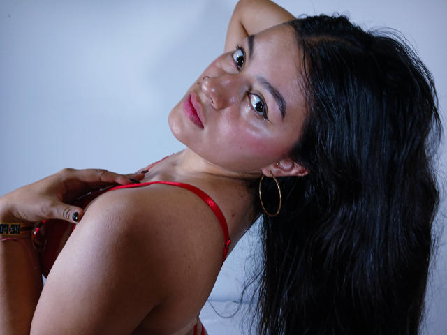 Catalina Navarro, con cabello negro largo, un vestido rojo con tirantes, aretes de aro dorados, un piercing en la nariz y una mano en el pecho, mirando por encima del hombro con una ligera sonrisa, mostrando sus senos..