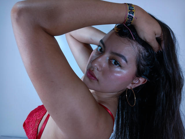 Catalina Navarro, con los brazos levantados, lleva un sujetador de encaje rojo, piel brillante, aros en las orejas, cabello negro, una pulsera y mira a la cámara con los labios ligeramente entreabiertos. Sus pechos son visibles..