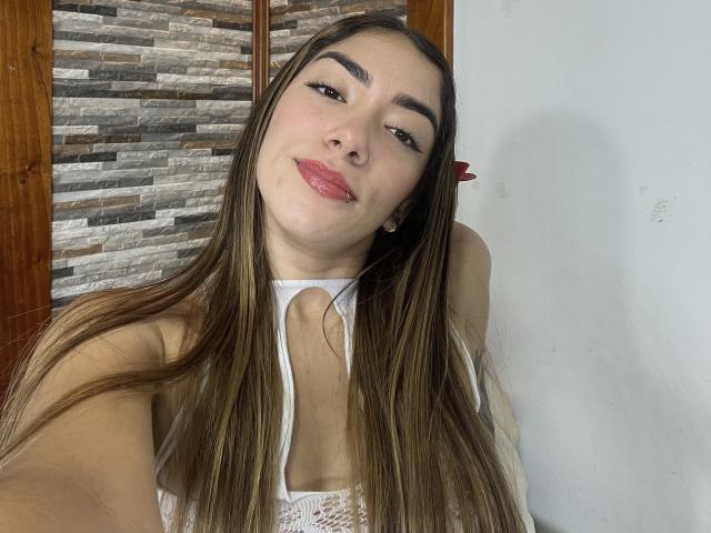 SharonKlart with long brown hair, white lace top, pink lipstick, and raised eyebrows, looking at the camera with a slight smile, showing her small tits.