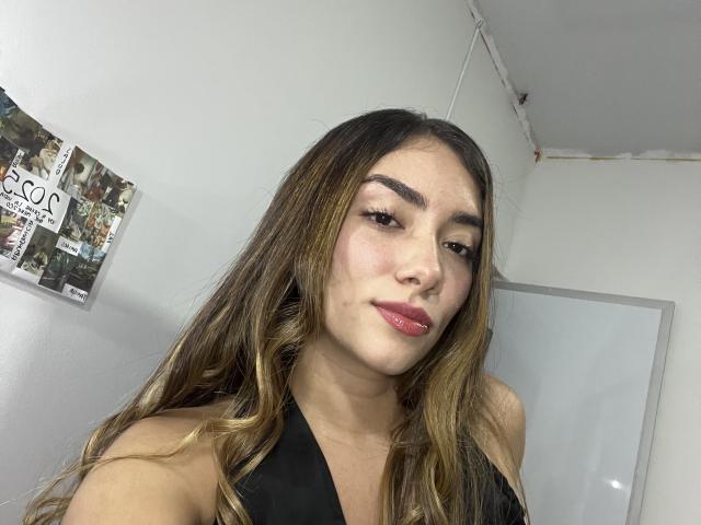 SharonKlart has long brown hair with blonde highlights, thick eyebrows, and red lipstick, wearing a black halter top, looking at the camera with a slight smile.