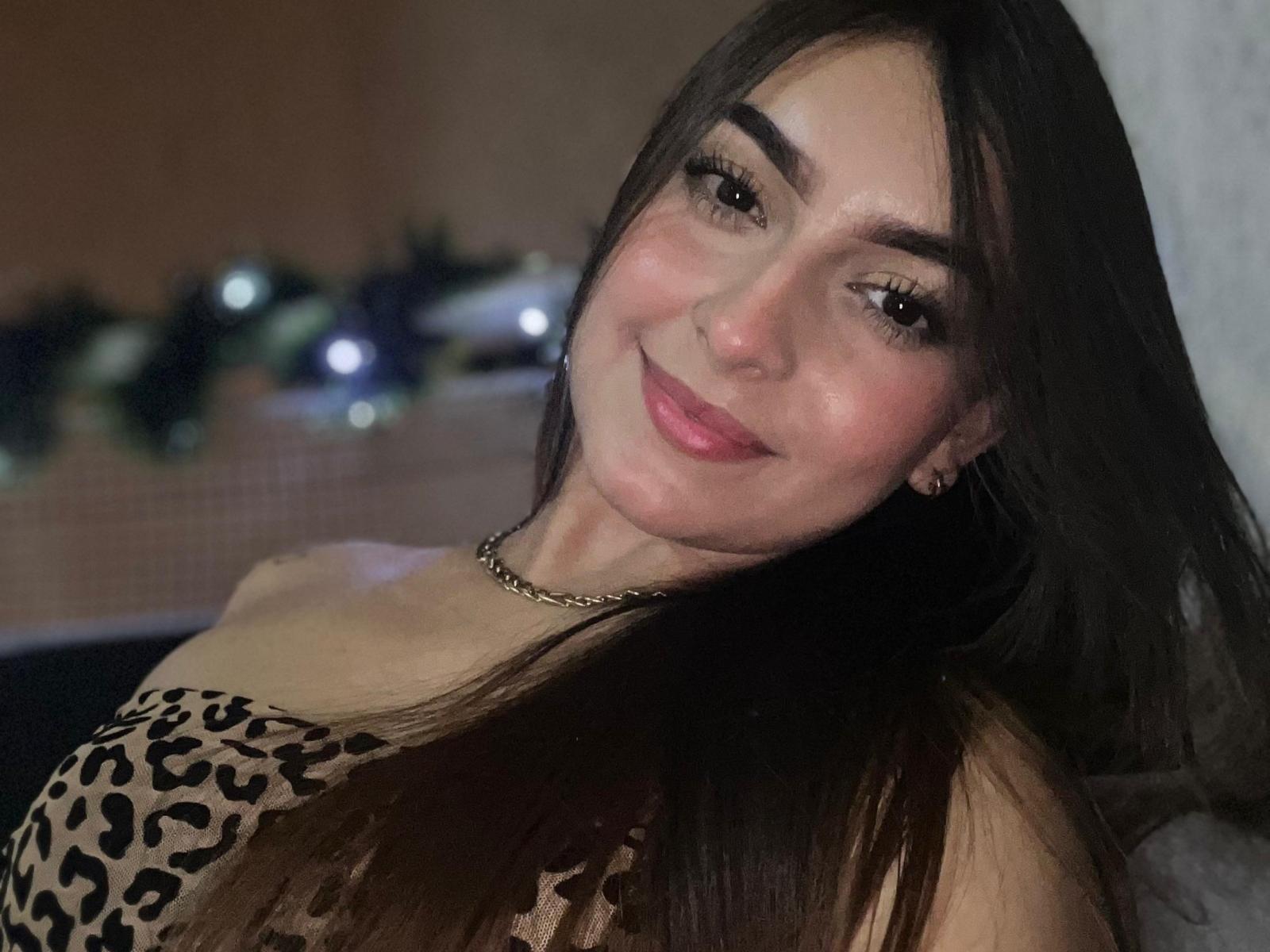 KarolTrue mit langen, braunen Haaren, die ein Top mit Leopardenmuster trägt, lächelt mit rosafarbenen Lippen, braunen Augen und einer goldenen Halskette und zeigt dabei ihre Brüste. Sie ist süß und sexy..