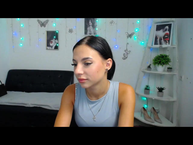 OliviaFoxy with dark hair, wearing a light gray sleeveless top, looking down, showing off her tits with a subtle makeup look, necklace, and blue fairy lights in the background.