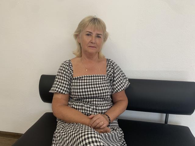 NoraVeda with blonde hair, wearing a black and white checkered dress, sitting on a black bench, hands on her lap, looking slightly to the side, wearing a bracelet.