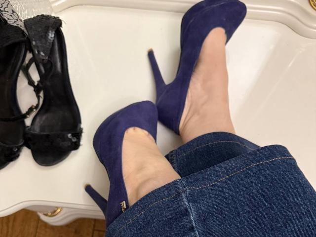 MiraGi's legs in denim jeans, wearing purple high heels, with black strappy sandals on a white surface. Visible toes, pale skin, wooden floor.