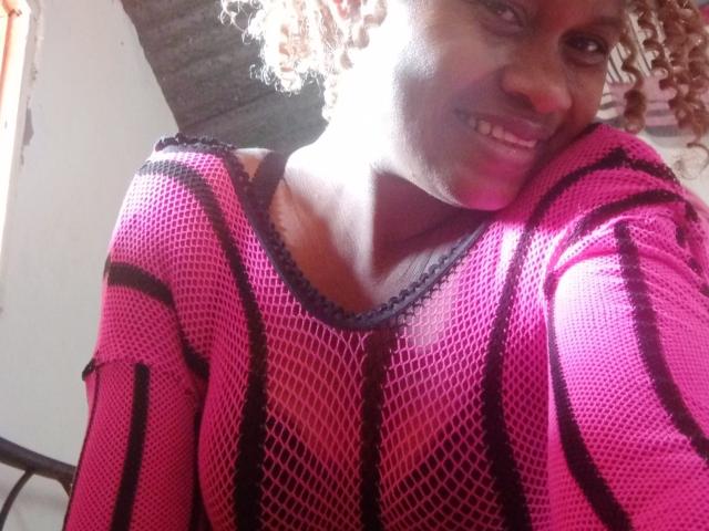 PrettySylvia, avec ses cheveux blonds bouclés, souriante, portant un haut en maille rose avec des rayures noires, laissant entrevoir ses seins sous la maille, debout dans une lumière du soleil vive, regardant l'appareil photo..