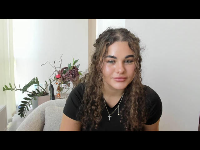 GloriaLi with curly brown hair, wearing a black shirt and cross necklace, smiling at the camera, sitting in a chair with a potted plant in the background.