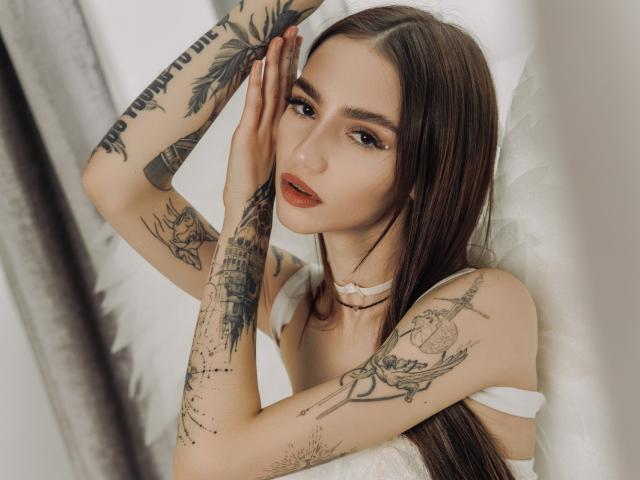 SophieKitty with long brown hair, tattoos on arms and chest, white strap dress, hands on face, red lipstick, looking at camera, white feather background, soft light.
