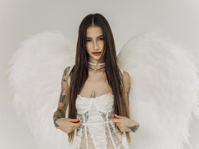 SophieKitty with long brown hair, white lace corset, large white angel wings, tattooed arms, choker, and red lipstick, holding corset straps, showing small white-boobs.