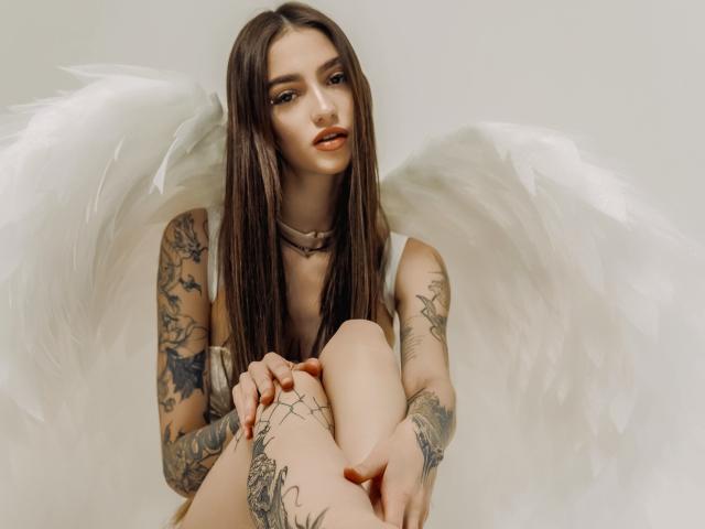 SophieKitty with long brown hair, tattoos on arms and legs, white angel wings, sitting with knees up, looking at camera, wearing a choker, neutral expression.
