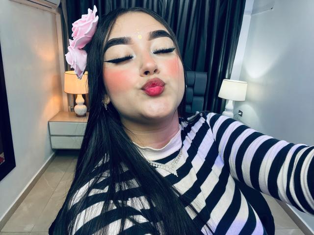 AnaShalimarr, puffy pink lips puckered, black striped shirt, long black hair with pink flower, eyeliner wings, red blush, eyes closed, white necklace, black earrings, small yellow dot on forehead.
