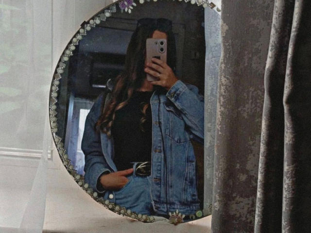 FlowerLeila-hot takes a mirror selfie wearing a blue denim jacket, black top, and high-waisted jeans, with long wavy brown hair, holding a smartphone in a dim room.