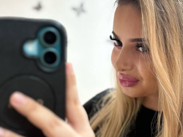 SinfullKitty with blonde hair taking a selfie, showing plump pink lips and long lashes, holding a black phone with a blue camera.
