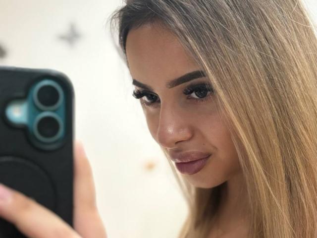 SinfullKitty with long blonde hair, full eyebrows, and purple lipstick, holding a black phone, taking a selfie, showing her cute face and slightly smiling.