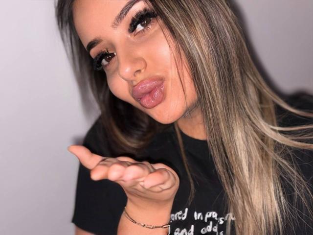 SinfullKitty with long, straight brown hair, pouty lips, and thick eyelashes, sticking out her tongue, pointing with her right hand, wearing a black shirt.
