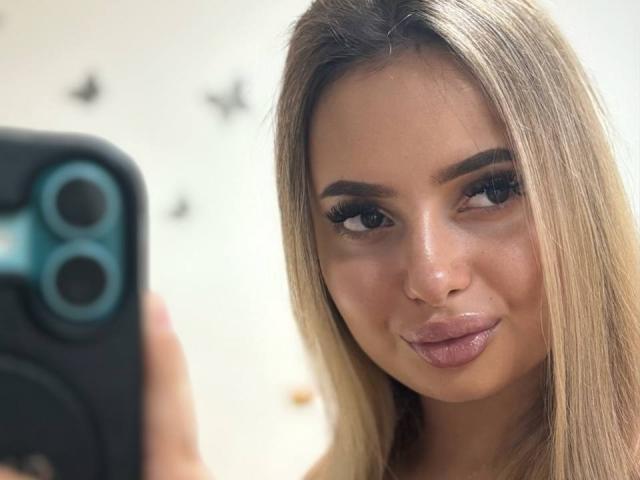 SinfullKitty has blonde hair, full pouty lips, and is taking a selfie with a black phone, showing off her thick, dark eyebrows and long lashes.