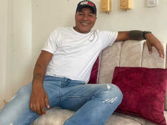 AxelStone, medium-skinned, smiling man with tattoos, wearing a white tee, blue jeans, and a cap, leans on a couch with a red pillow, right arm resting on the back.
