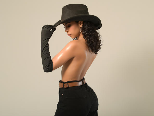 JadeMiranda with curly brown hair, wearing a black hat, black gloves, and black high-waisted pants, topless, showing her shiny bare back and ass, holding her hat.