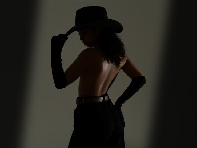 Silhouetted JadeMiranda with curly hair, wearing black gloves, hat, and pants, back to camera, topless, showing her ass, holding hat with right hand, dark shadowy background.