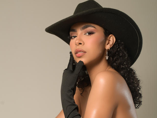 JadeMiranda with curly black hair, wearing a black hat and gloves, touching her cheek with her gloved hand, showing her shiny bare tits and a confident, sexy expression.