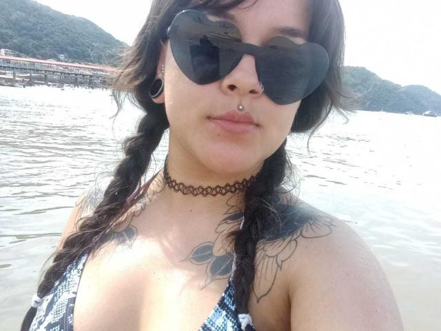 DaisyFx with braided hair, black sunglasses, nose piercing, choker, tattoos on shoulders, and a blue checkered bikini, standing in water, showing her tits.