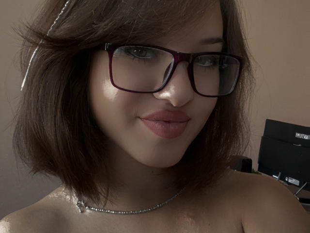 Ssimona with dark brown bob, wearing red-framed glasses, shiny pink lipstick, and a silver necklace, showing a subtle smile, no clothes visible, fair skin, and a relaxed look.
