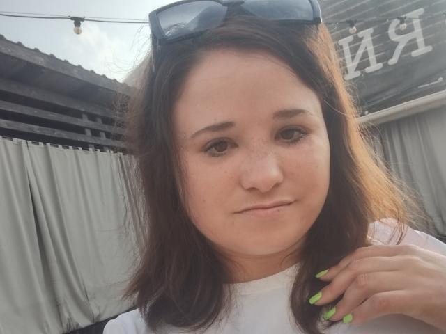 Annoying has light skin, brown eyes, and brown hair with green nails, wearing sunglasses on her head, and a white shirt, looking slightly annoyed with a subtle smile.