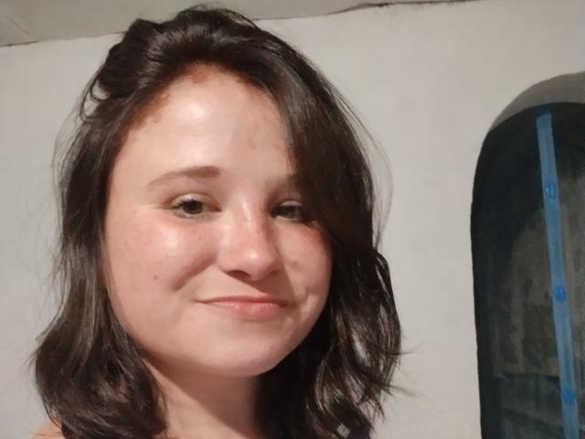 Annoying woman with medium-length, wavy brown hair, slightly flushed cheeks, small nose, and a subtle smile. She's looking at the camera with a relaxed, slightly cute expression.
