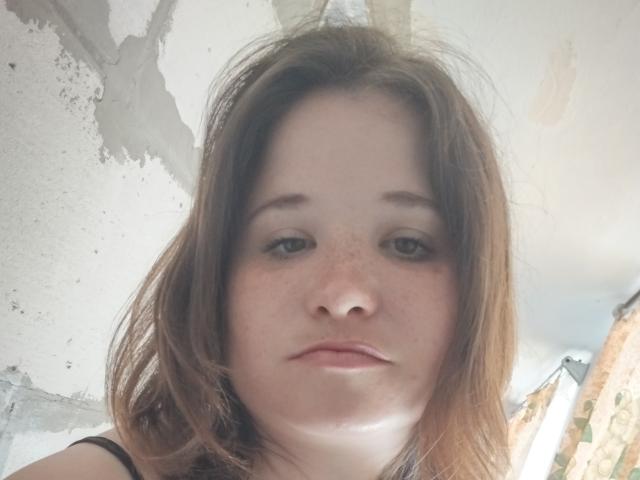 Annoying has light brown, slightly messy hair, pale skin with freckles, and is looking down with a neutral expression. Her small nose and subtle pink lips are visible.
