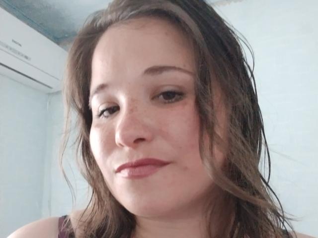 Annoying woman with brown hair, freckles, and pink lipstick, wearing a purple tank top, has a slightly annoyed expression, and is taking a selfie.
