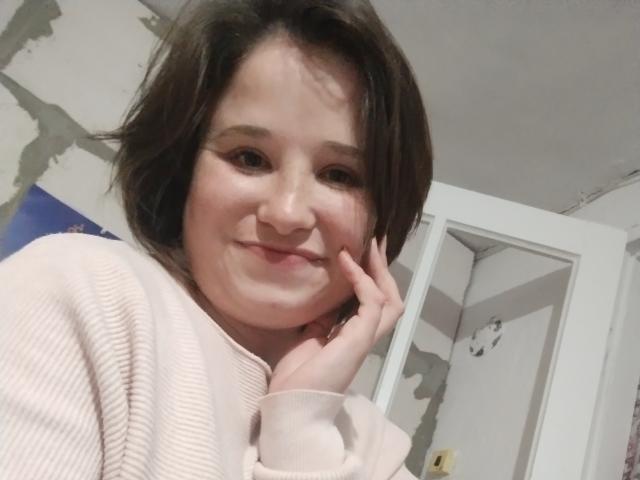 Annoying with short brown hair, wearing a pink ribbed sweater, smiling slightly, touching her cheek with her left hand, showing subtle makeup and a cute face.